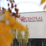 The background is in focus - there is a white building that reads "Central Baptist Theological Seminary." In the foreground is blurry orange flowers or leaves.
