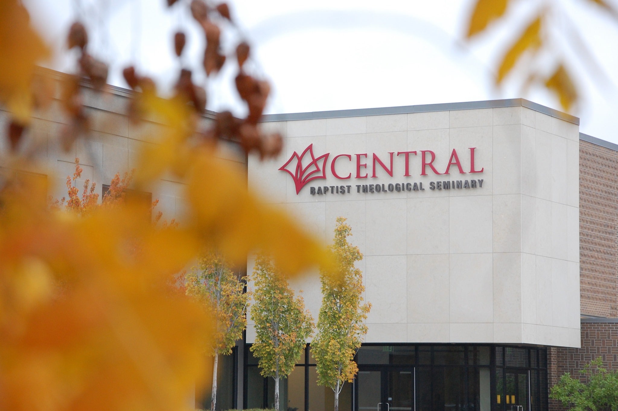 The background is in focus - there is a white building that reads "Central Baptist Theological Seminary." In the foreground is blurry orange flowers or leaves.