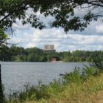 Image of a lake surrounded by grasses, trees, and other plants. In the background is a light brown building.