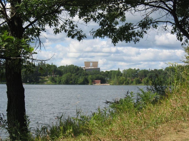 Image of a lake surrounded by grasses, trees, and other plants. In the background is a light brown building.
