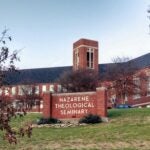 Image of a long red brick building with a black roof. Red and brown trees are scattered through the picture. Centered is a red brick sign with white letters that reads "Nazarene Theological Seminary."