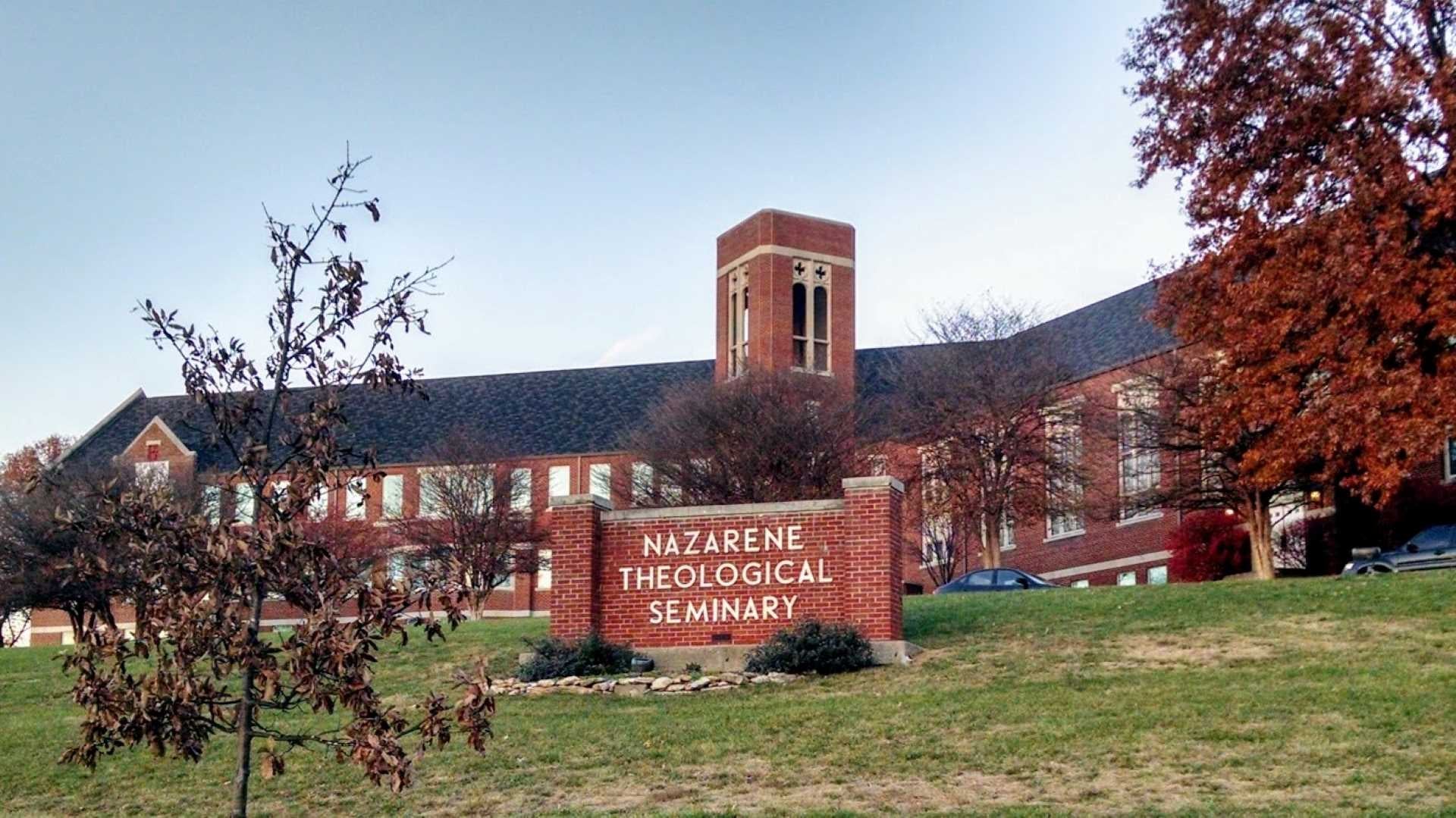 Image of a long red brick building with a black roof. Red and brown trees are scattered through the picture. Centered is a red brick sign with white letters that reads "Nazarene Theological Seminary."