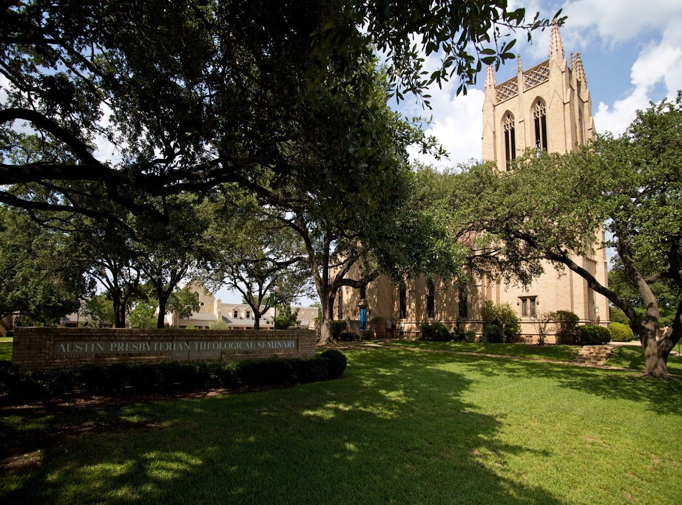 Image of green trees and grass that is partially in the shade, partially in the sun. In the right background is a light brown brick building with spires. To the left is a sign under the shade that reads "Austin Presbyterian Theological Seminary."