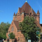 Image of a red-brown brick building with spires surrounded by green trees and a very blue sky.